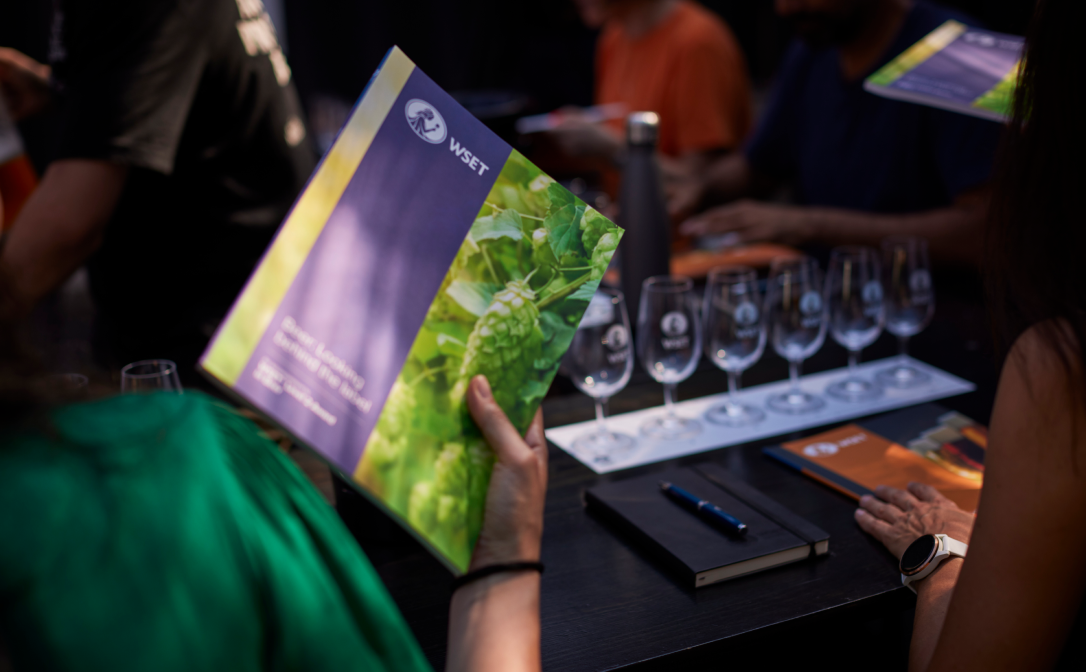 A person holding the WSET Level 2 Award in Beer textbook, with a row of glasses in the background